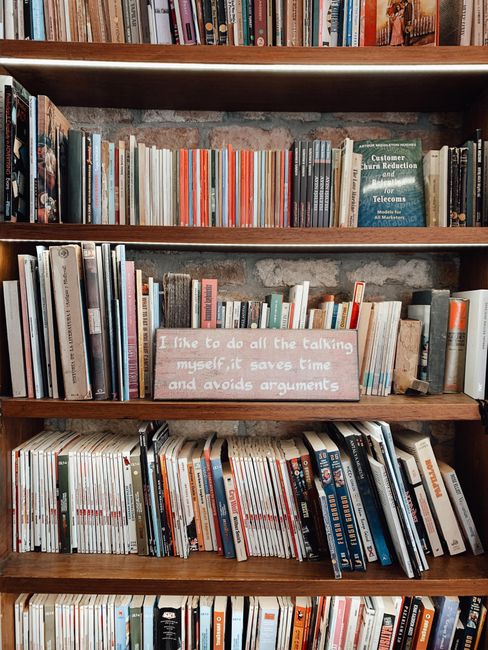A wooden bookshelf filled with a variety of books of different sizes and colors, with some leaning against each other. The middle shelf features a pink sign with white text that reads, 'I like to do all the talking myself, it saves time and avoids arguments.' The background shows a textured wall, and the lighting highlights the warm tone of the wooden shelves and the collection of books organized in a casual manner.